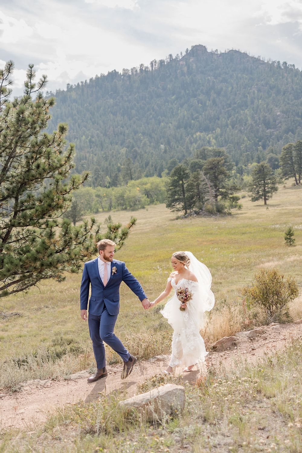 A couple in wedding attire dances playfully on a hillside with mountains and pine trees in the background.