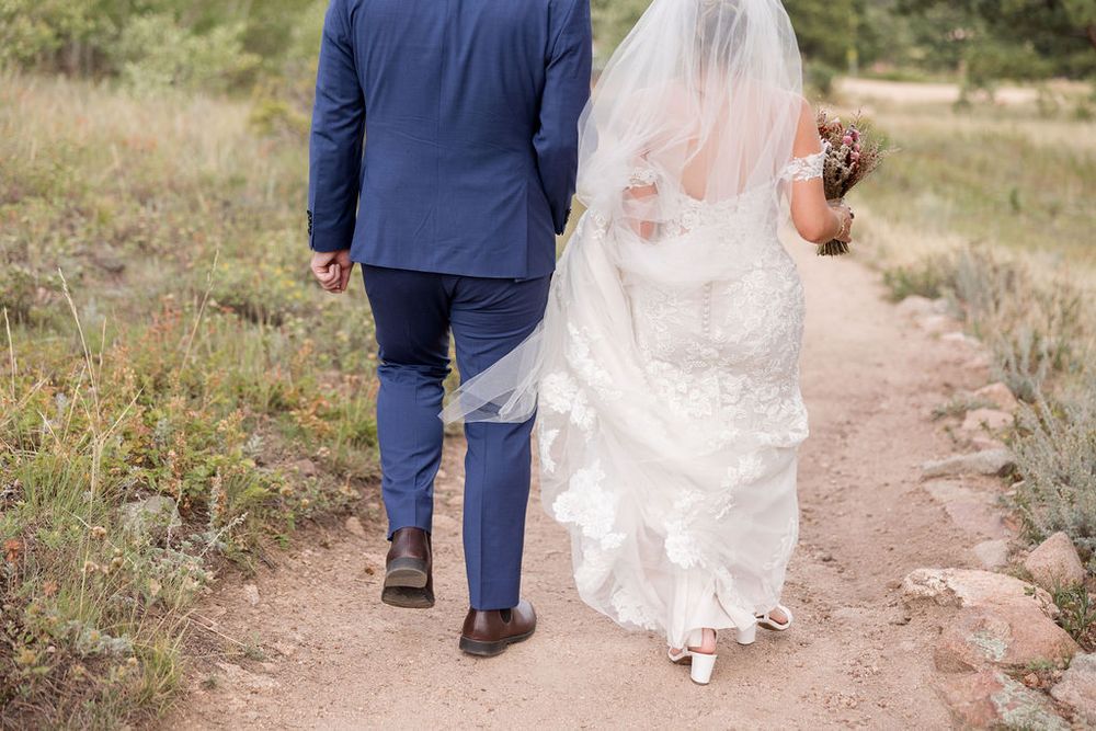 Wedding couple walking hand in hand down a rustic dirt path surrounded by mountain scenery.