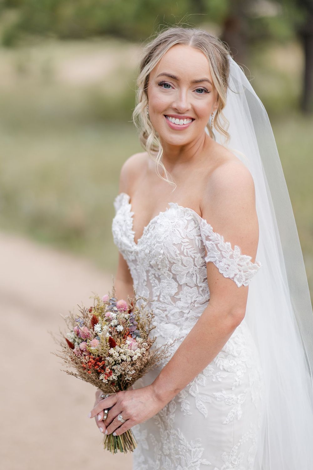 A bride in a lace wedding dress with off-shoulder detail holds a rustic bouquet of dried flowers and blush blooms.