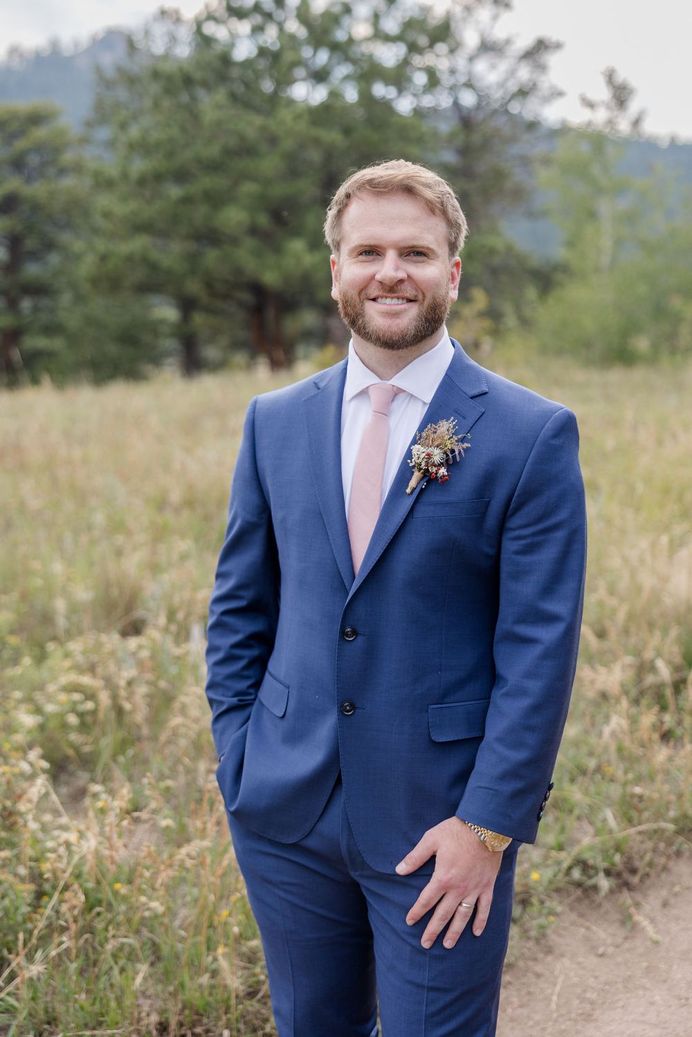 Groom in navy blue suit with pink tie poses outdoors in a field with mountains and trees in the background.