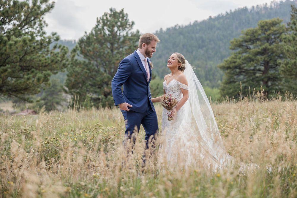 Newlyweds share a romantic moment in a mountain field surrounded by pine trees and golden grass.