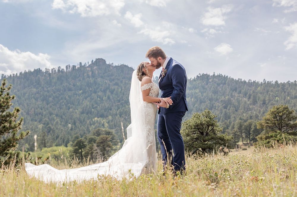 A couple in wedding attire stands together on a scenic mountain hillside with evergreen trees in the background.