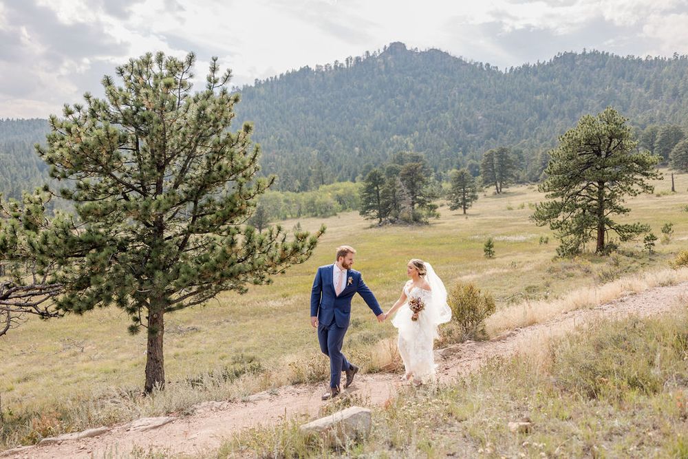 A couple in wedding attire walks through a scenic mountain meadow with tall grass and evergreen trees.