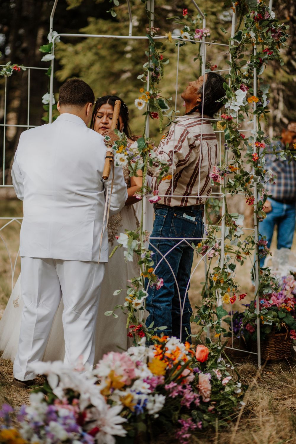 Guests in white attire gather at an outdoor wedding ceremony decorated with colorful floral arrangements and vines.