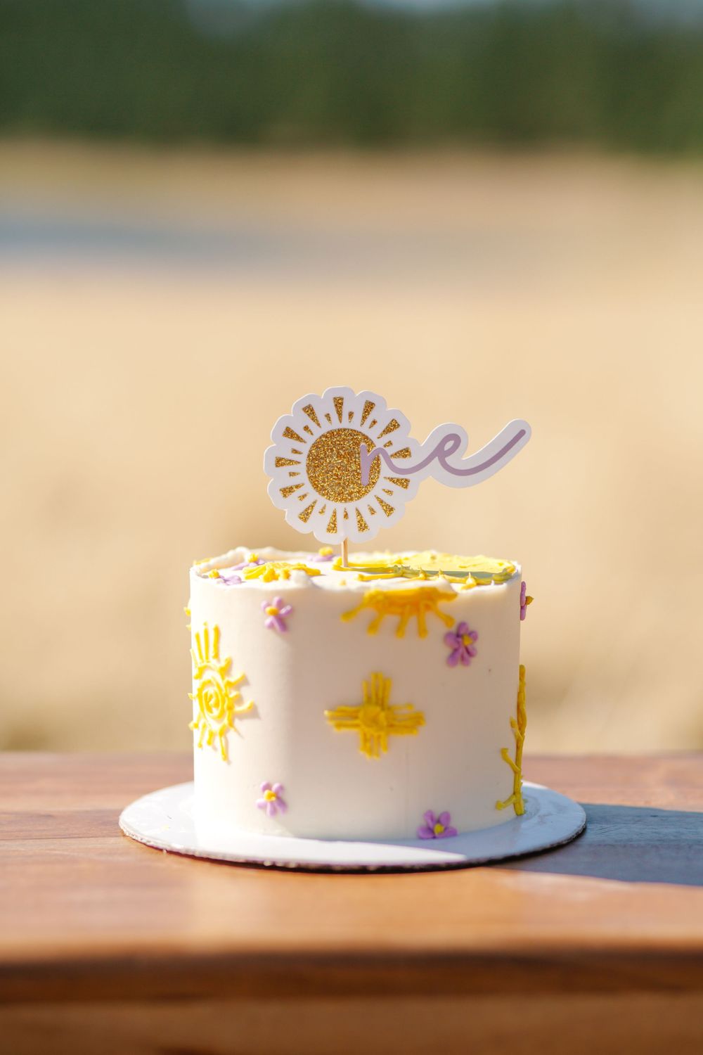 White frosted cake with yellow sun decorations and purple accents sits on wooden surface in natural lighting.