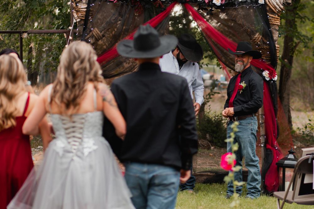 Wedding ceremony guests stand in a garden setting with red and black decor elements visible in the background.