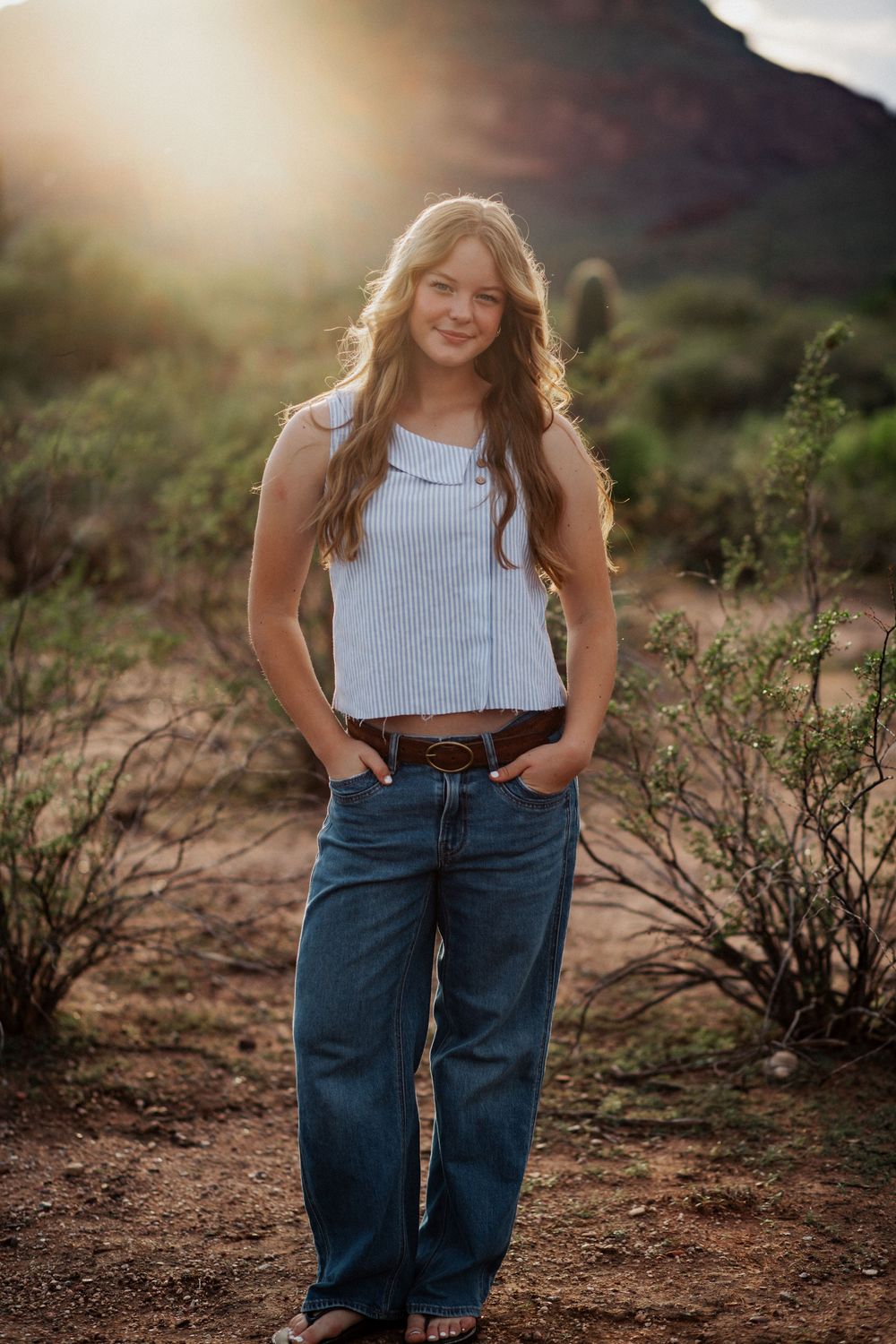 A person in a light blue crop top and relaxed fit jeans poses outdoors against a natural desert landscape at sunset.