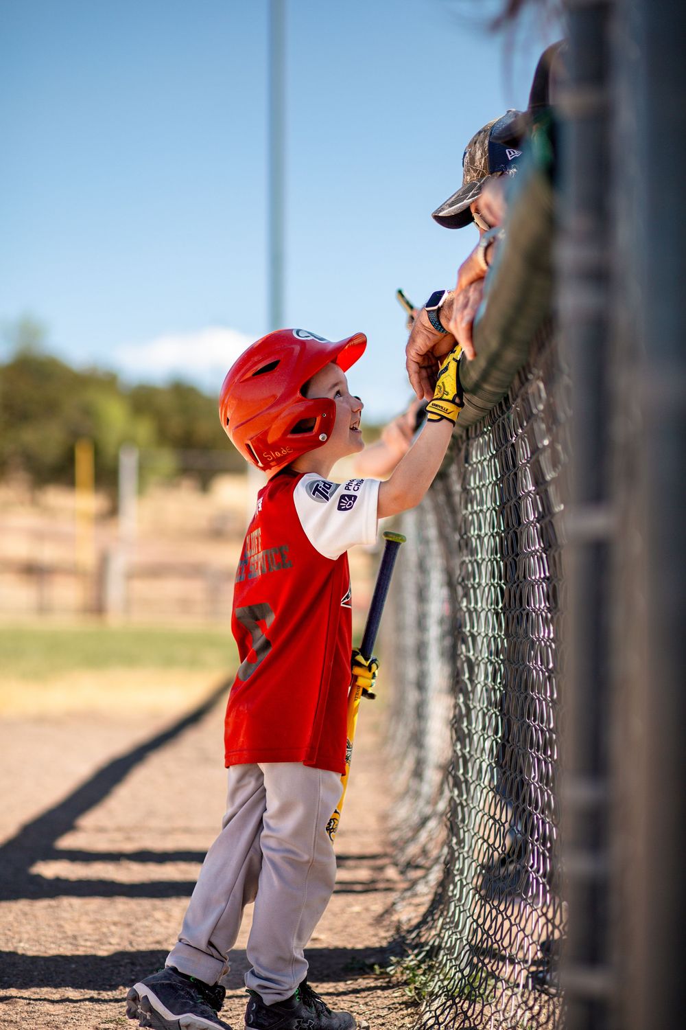 Young baseball player in red jersey and batting helmet reaches up to grab a water bottle through chain link fence at dugout.