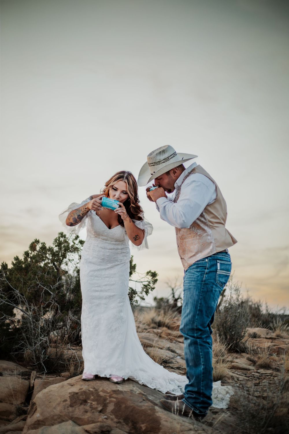 A couple in western attire shares a romantic dance pose against a desert landscape at sunset.