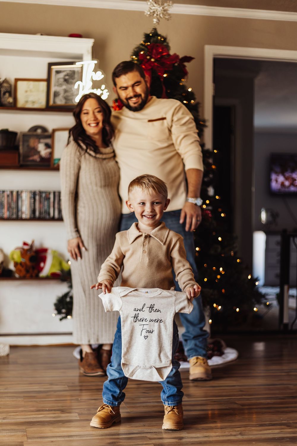 A toddler runs excitedly across hardwood floors with a paper bag while adults watch near a Christmas tree.