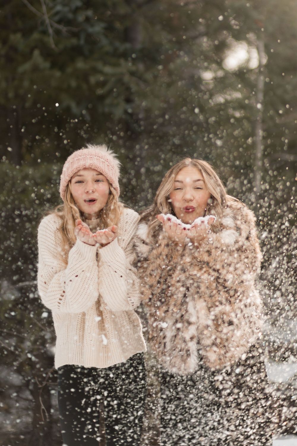 Two people in winter sweaters and knit hats playfully blow snow into the air during a snowy day outdoors.