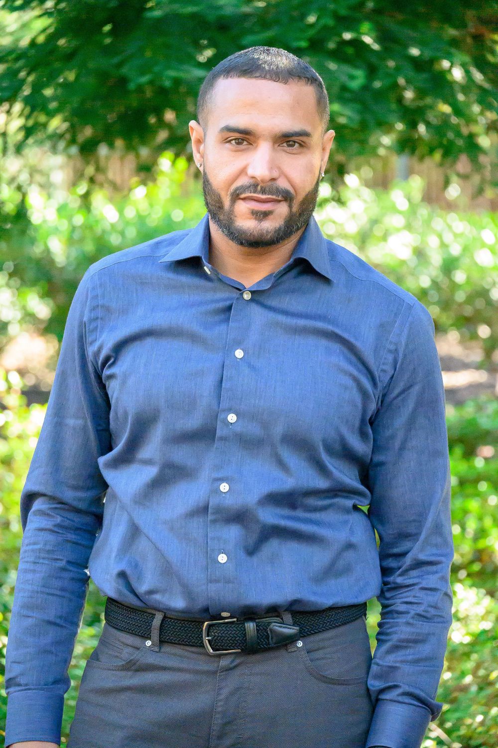 A person wearing a navy blue button-up shirt poses outdoors against a leafy green background.