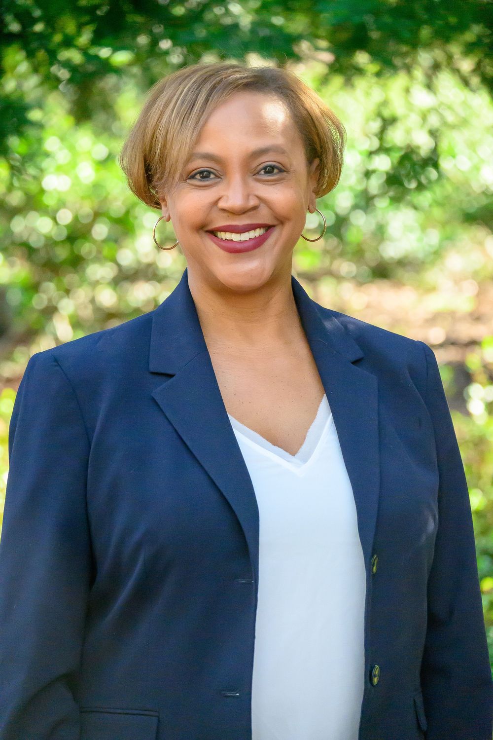 Professional wearing a navy blazer and white top stands in front of lush green foliage outdoors.
