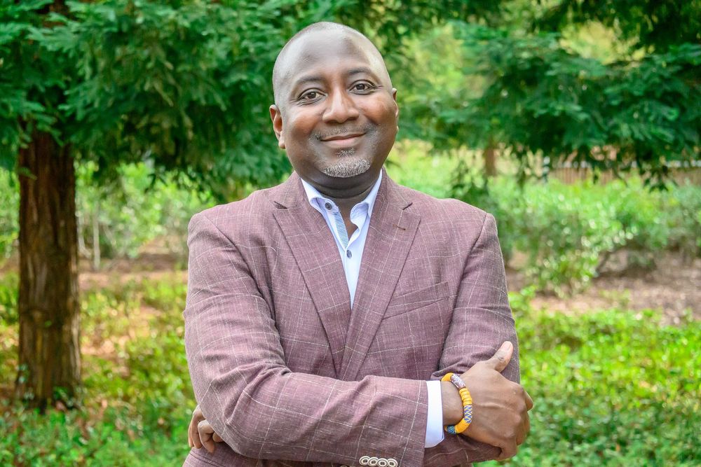 Professional portrait of someone in a brown blazer and blue shirt smiling warmly outdoors with green foliage in background.