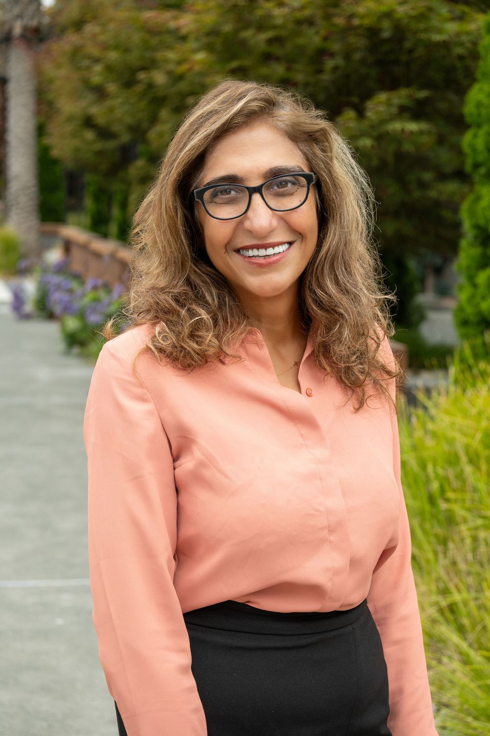 A person wearing glasses and a peach-colored top with black dress smiles warmly in an outdoor garden setting.