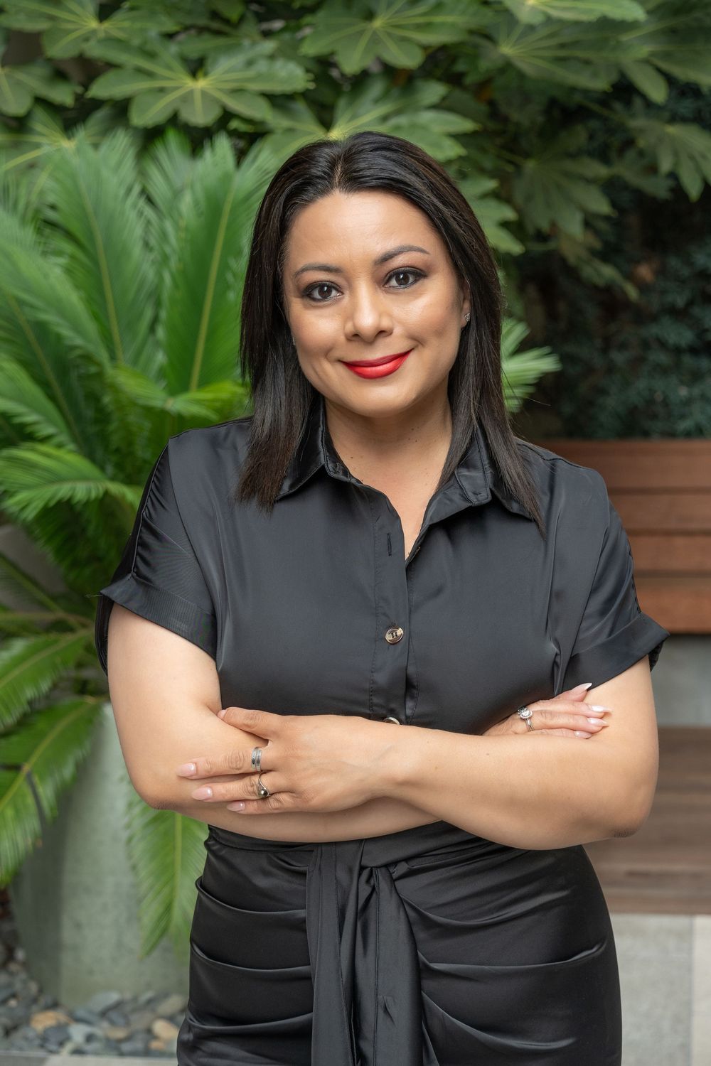 Professional portrait of someone wearing a black button-up shirt posing with crossed arms against a green garden background.