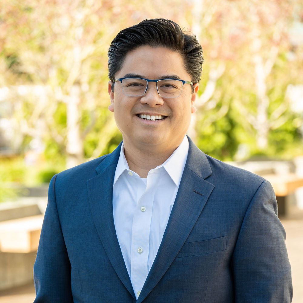 Professional portrait photo of a person in a navy suit and white shirt smiling outdoors with natural lighting.