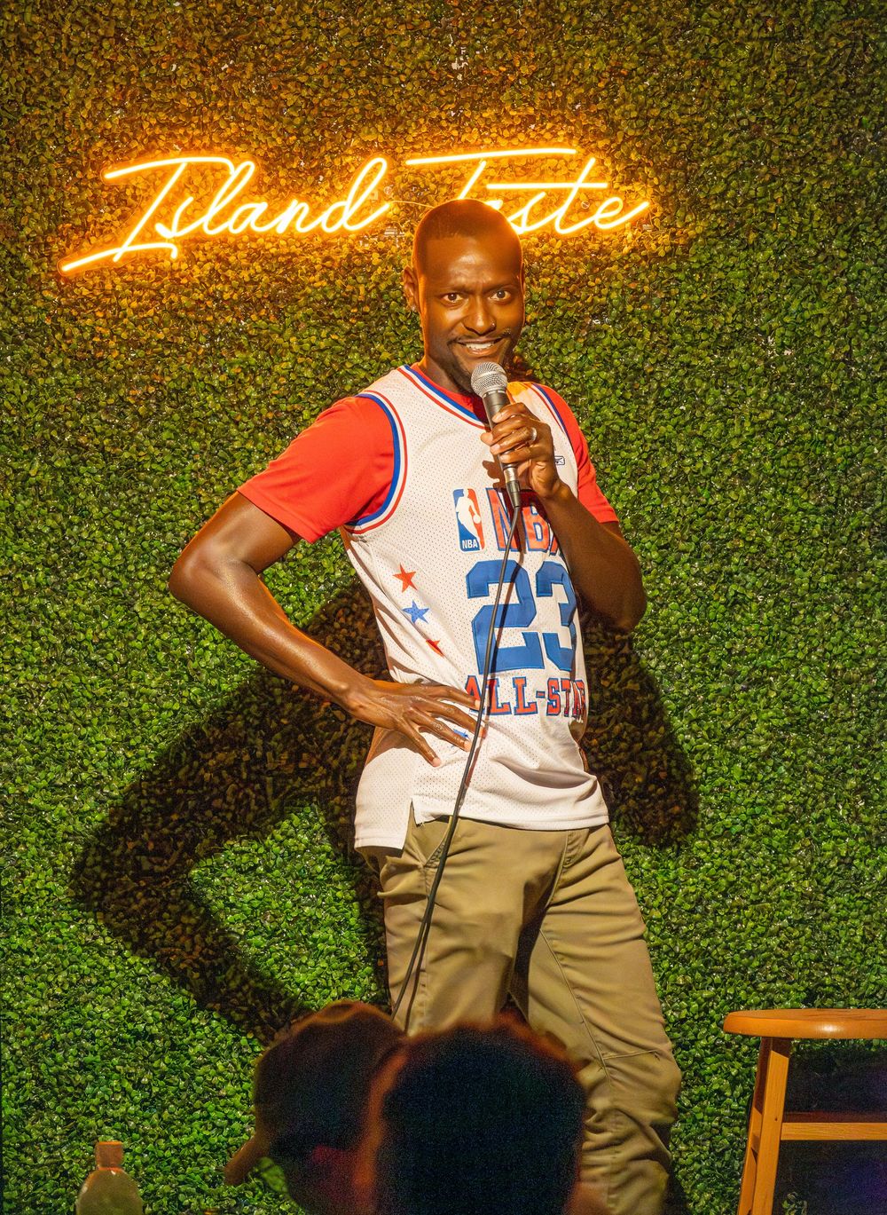 Comedian in basketball jersey performs stand-up against green wall with neon signage.