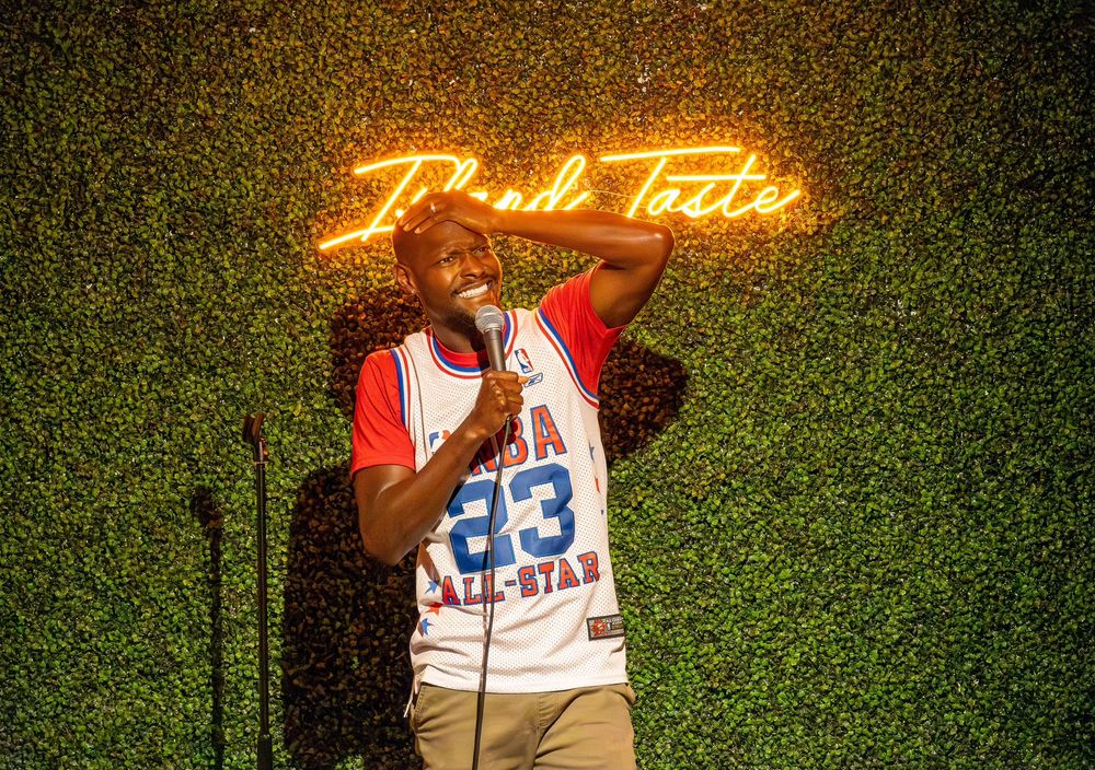 Performer in sports jersey delivers comedy routine on stage with neon lighting.