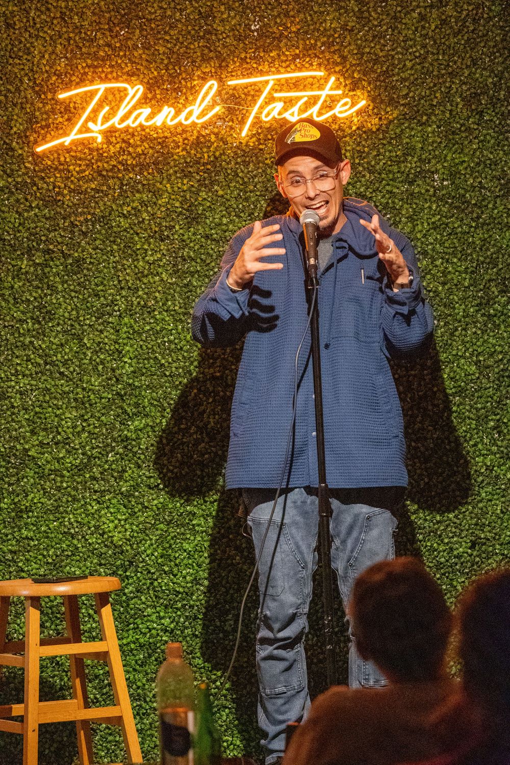 Comedian performing stand-up routine on stage with neon sign and green foliage backdrop.