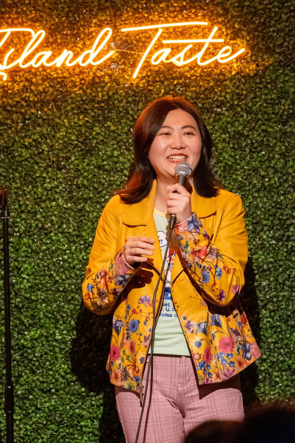 Performer in yellow floral jacket delivers comedy set against illuminated green wall backdrop.