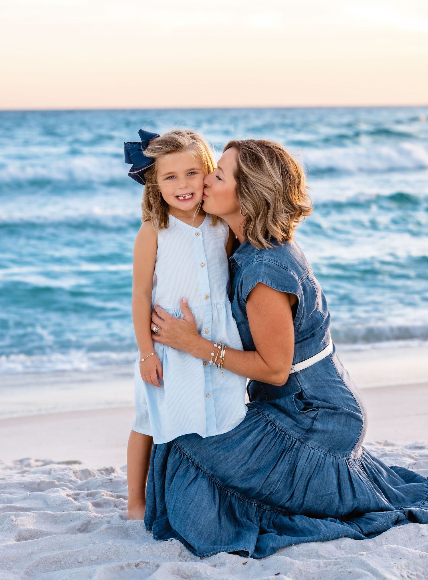A tender beach scene with figures in blue and white outfits embracing on white sand with ocean waves in background.