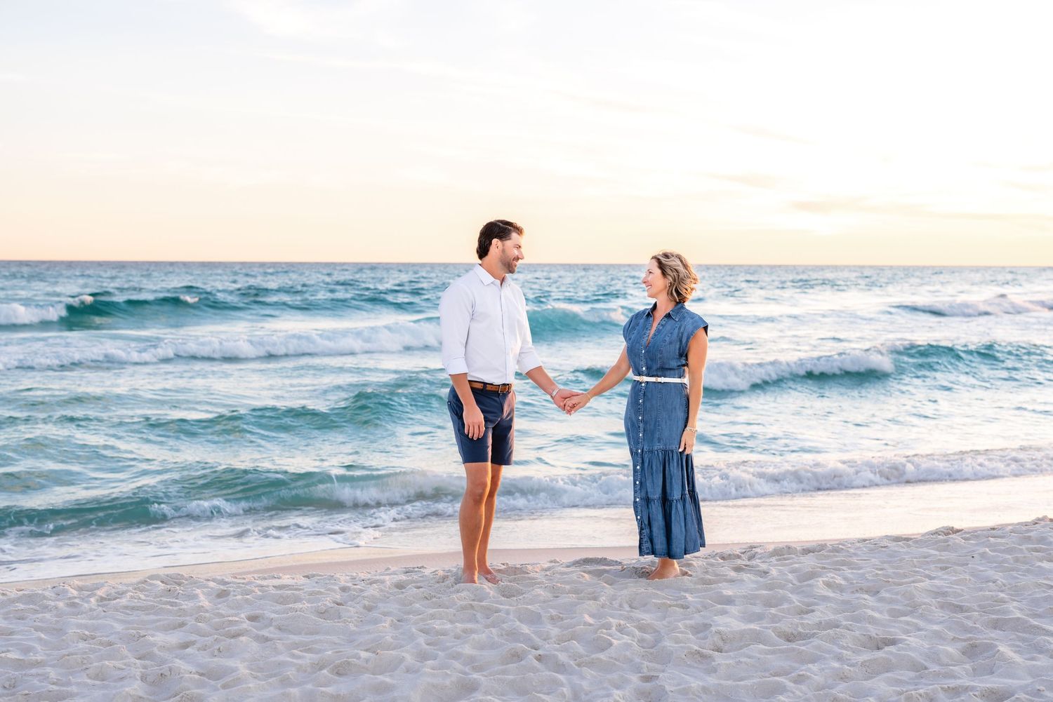 A couple walks hand in hand along a serene beach at sunset with waves rolling in along the shoreline.