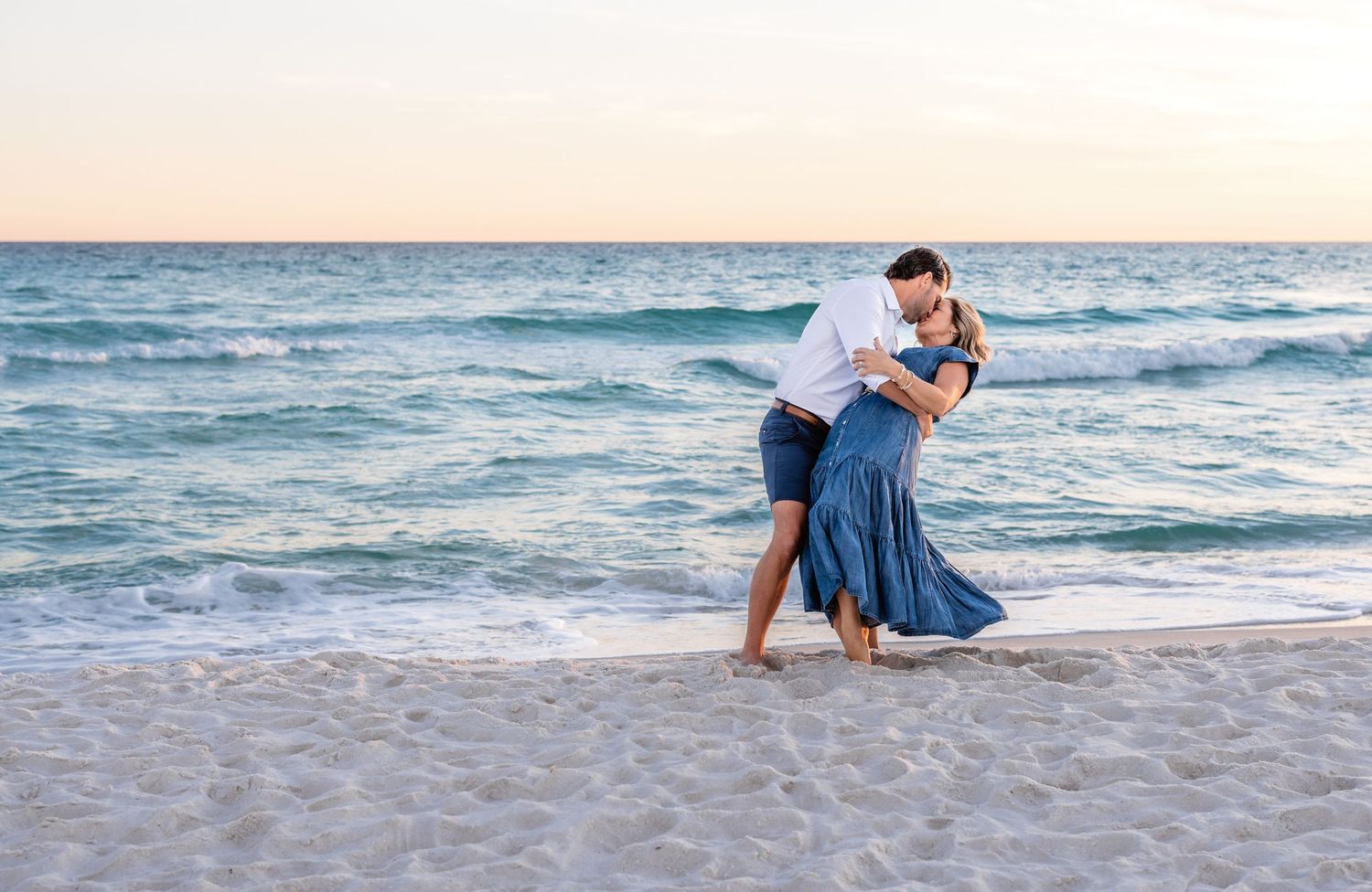 A couple in blue outfits poses romantically together on a sandy beach at sunset.