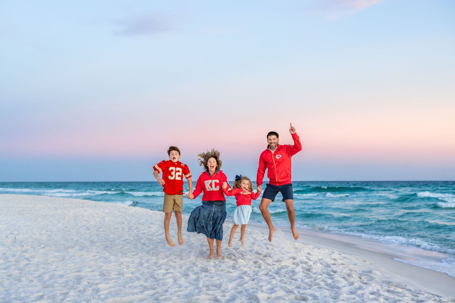 Family wearing red shirts poses together on white sandy beach at sunset with ocean waves in background.