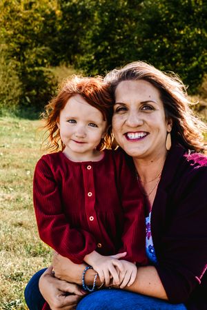 A parent and child sit together outdoors in autumn sunlight wearing red and dark clothing against a green background.