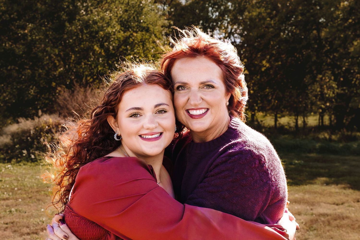 Two people in red and purple sweaters embrace while smiling outdoors on a sunny autumn day.