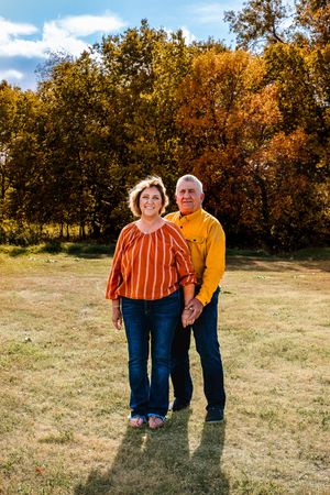 A couple stands together in a field during autumn with golden trees and blue sky in the background.