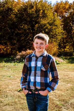 A child wearing a plaid shirt and jeans stands outdoors in autumn with golden foliage in the background.