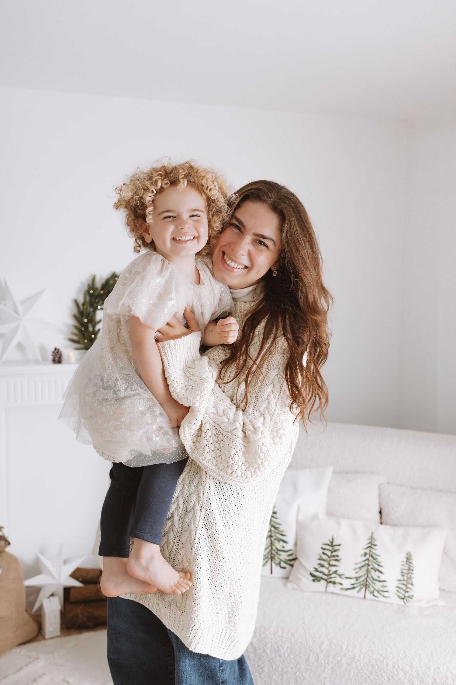 Mother and child in matching white sweaters share joyful moments in a bright minimalist room with holiday decorations.