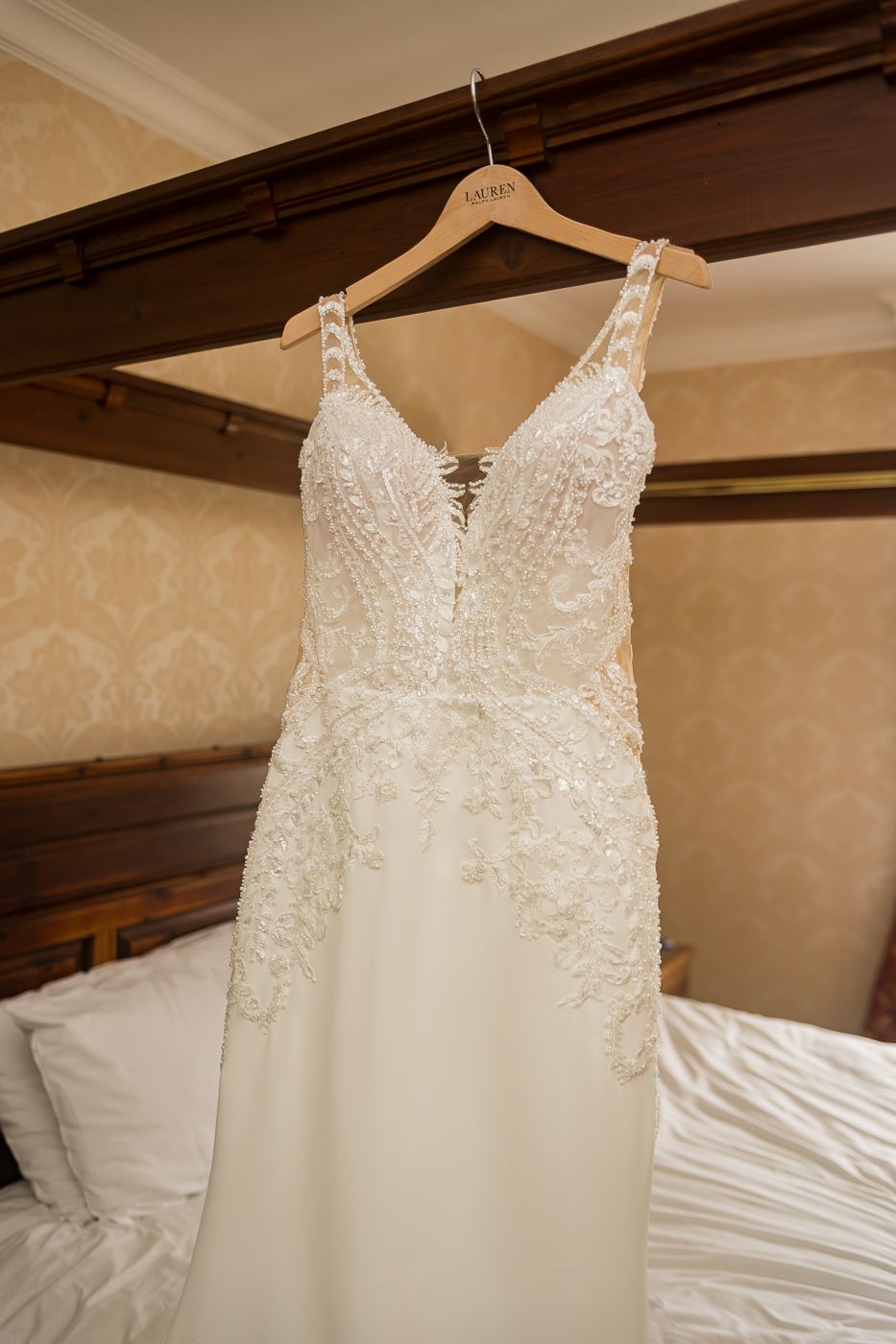 Elegant white lace wedding dress hanging on display against a beige wall.