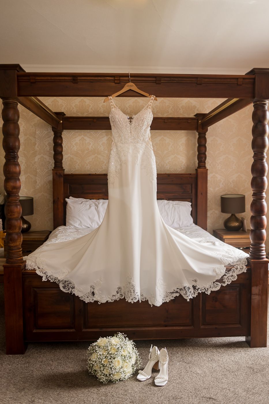 White wedding dress displayed with baby's breath bouquet and bridal shoes on carpet.