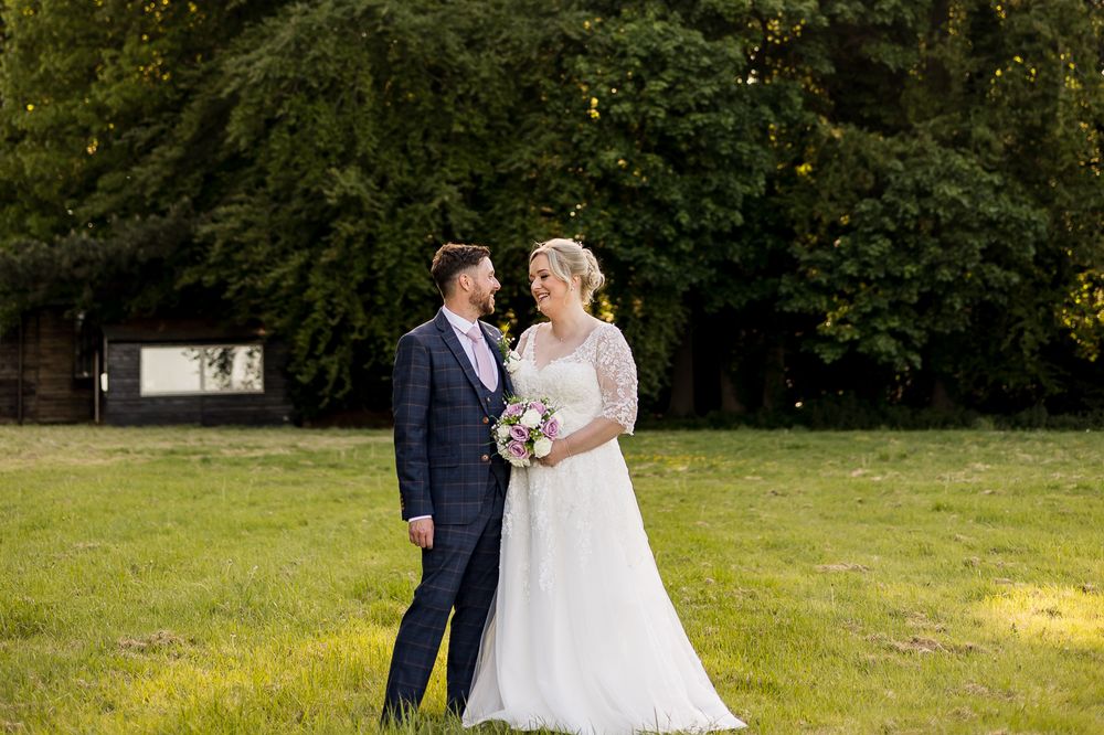Wedding couple stands together on grassy field with trees in background during sunset.