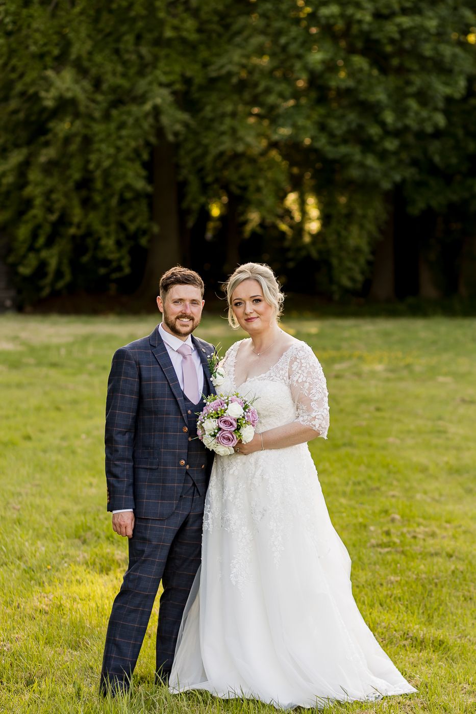 Wedding portrait of couple in formal attire standing in sunlit garden setting.