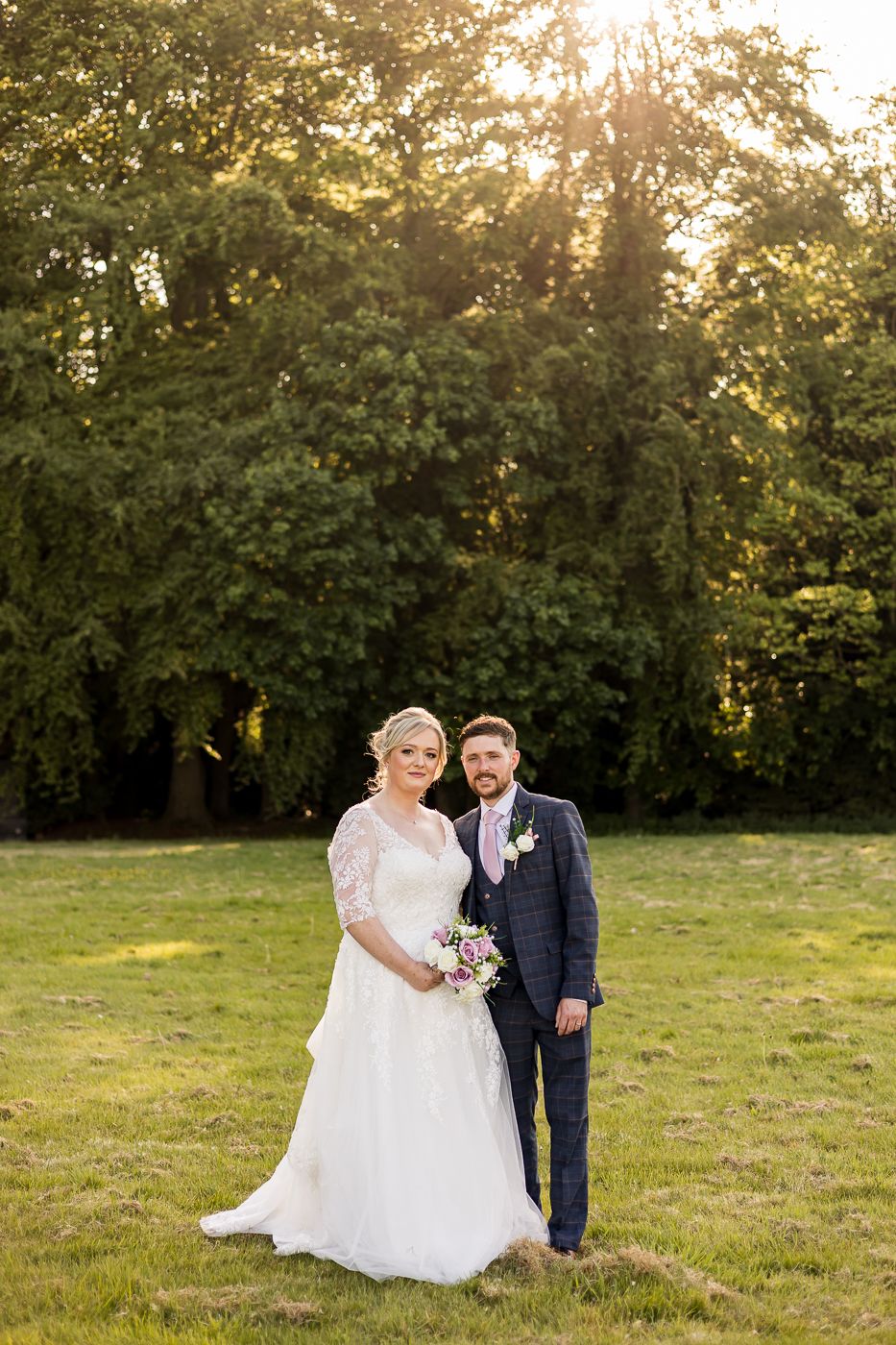 Newlyweds share a romantic moment in a sunlit meadow with trees in the background.