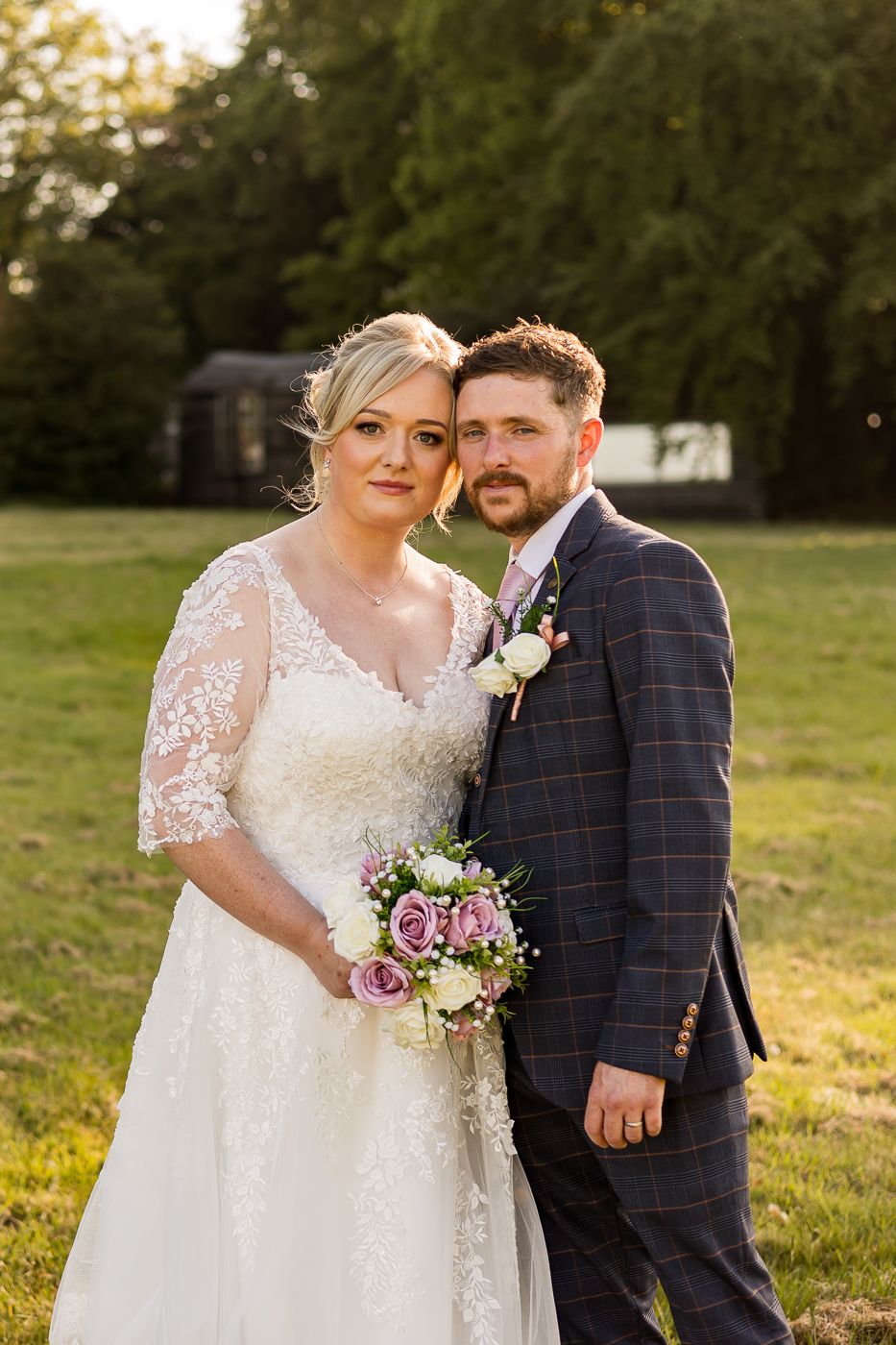Bride in lace dress and groom in plaid suit embrace while holding a purple and white bouquet.