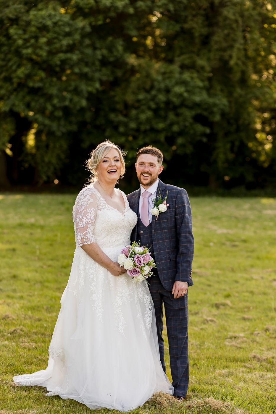 Joyful wedding couple laughs together on a grassy field during sunset.