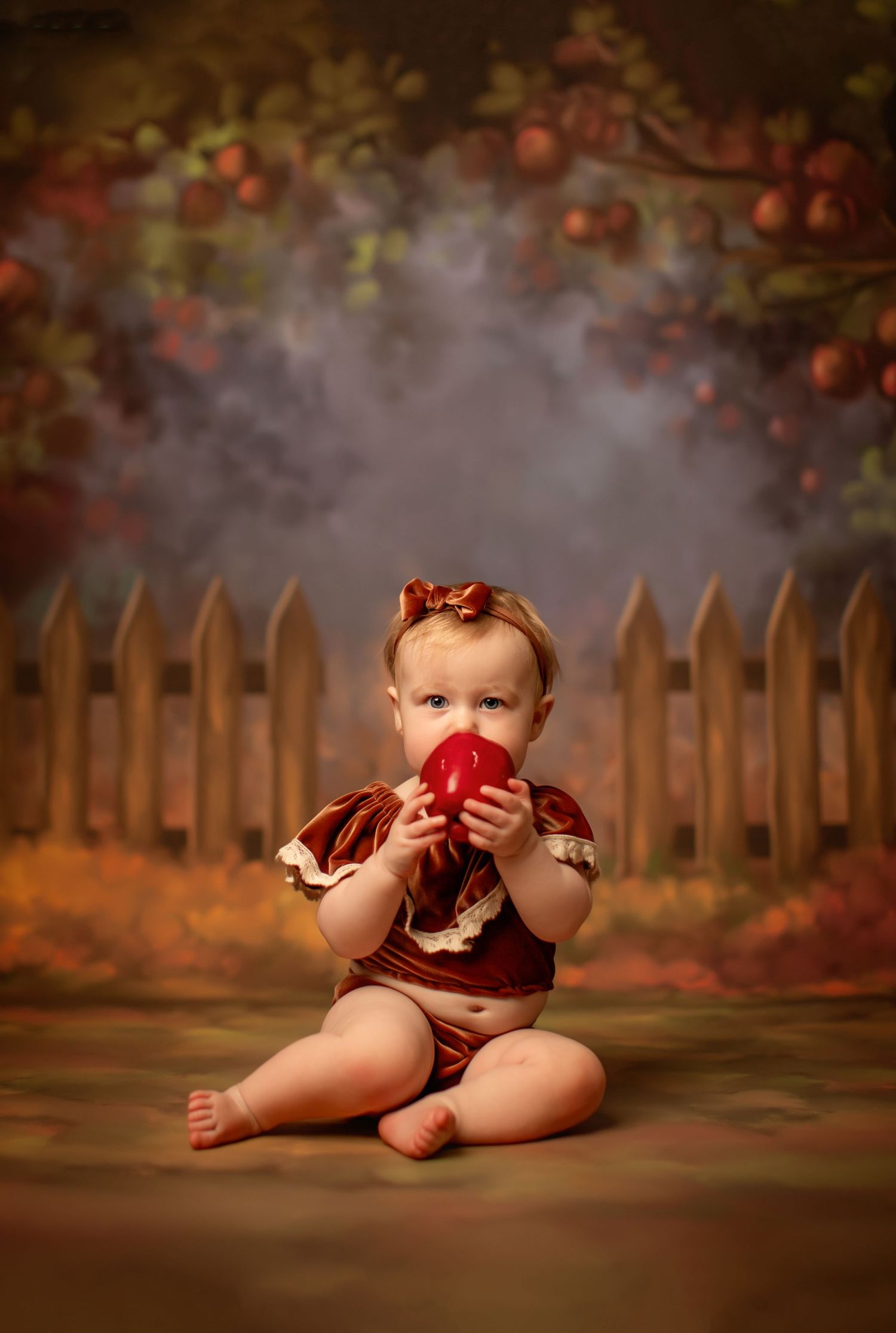 A baby dressed in burgundy sits on a dark floor with autumn leaves and a wooden fence in the background.