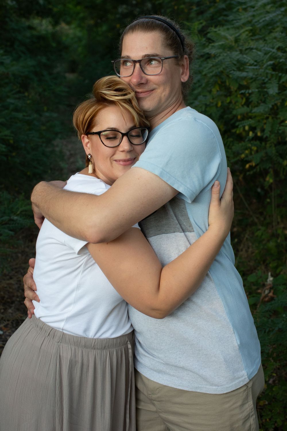 Two people share an intimate hug outdoors against a backdrop of green foliage.