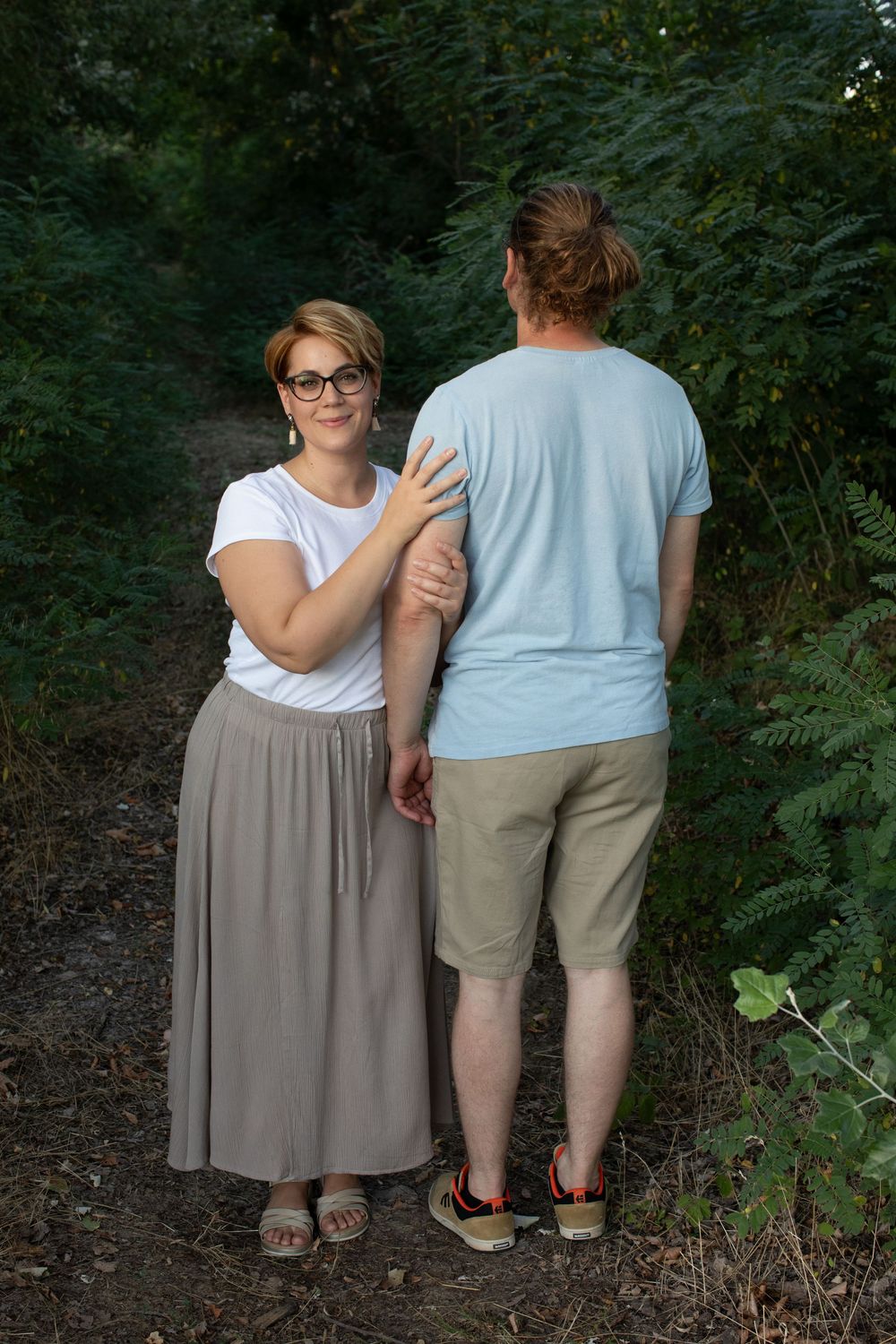Two people in casual summer attire walking together on a wooded trail.