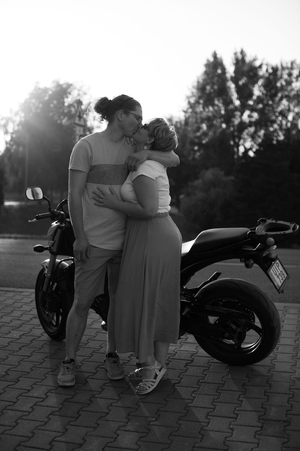 Silhouetted couple embracing near a parked motorcycle at sunset with trees in the background.