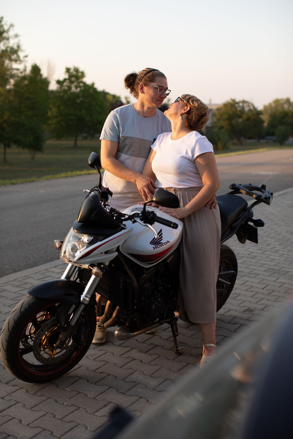 A couple poses together near a parked motorcycle on a paved outdoor area at sunset.