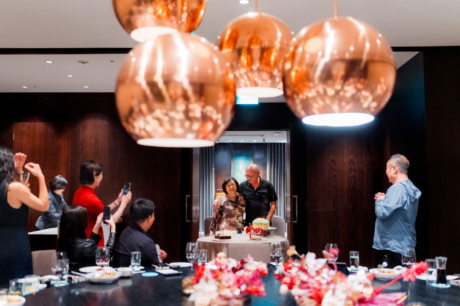 A dimly lit restaurant interior with copper pendant lights and diners seated at tables with red decor elements.