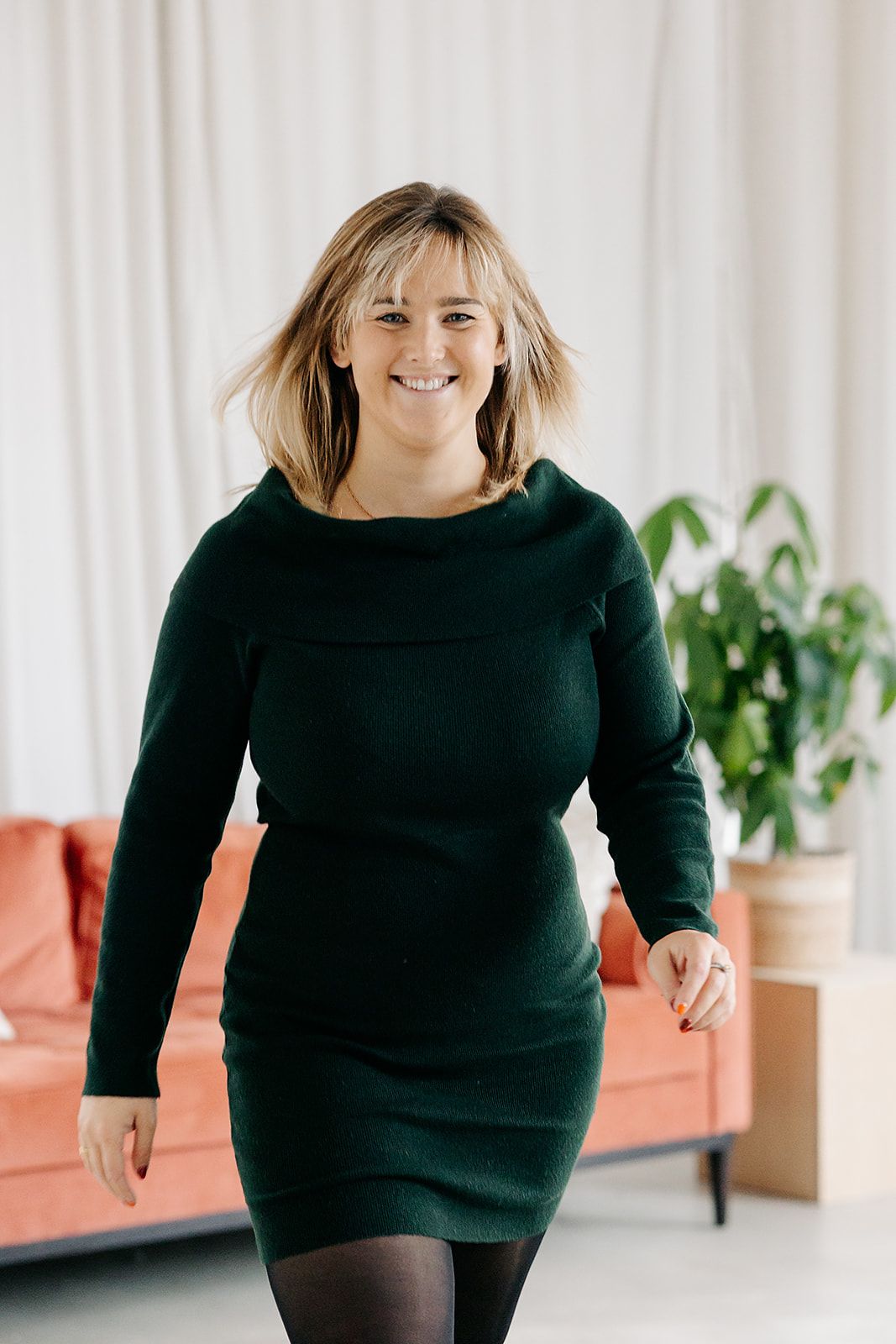 Woman in black sweater poses in bright interior space with houseplants and white curtains.