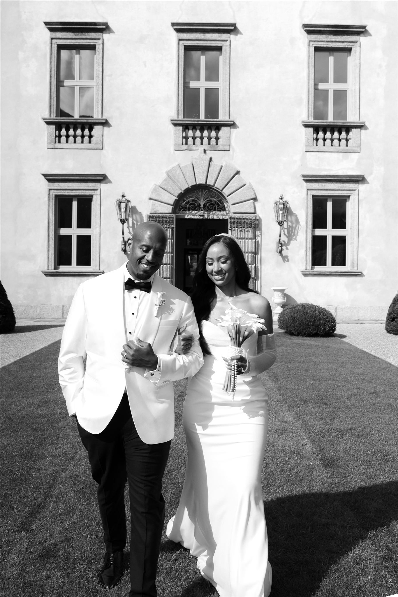 A wedding couple poses in front of an elegant white villa with classic architecture.