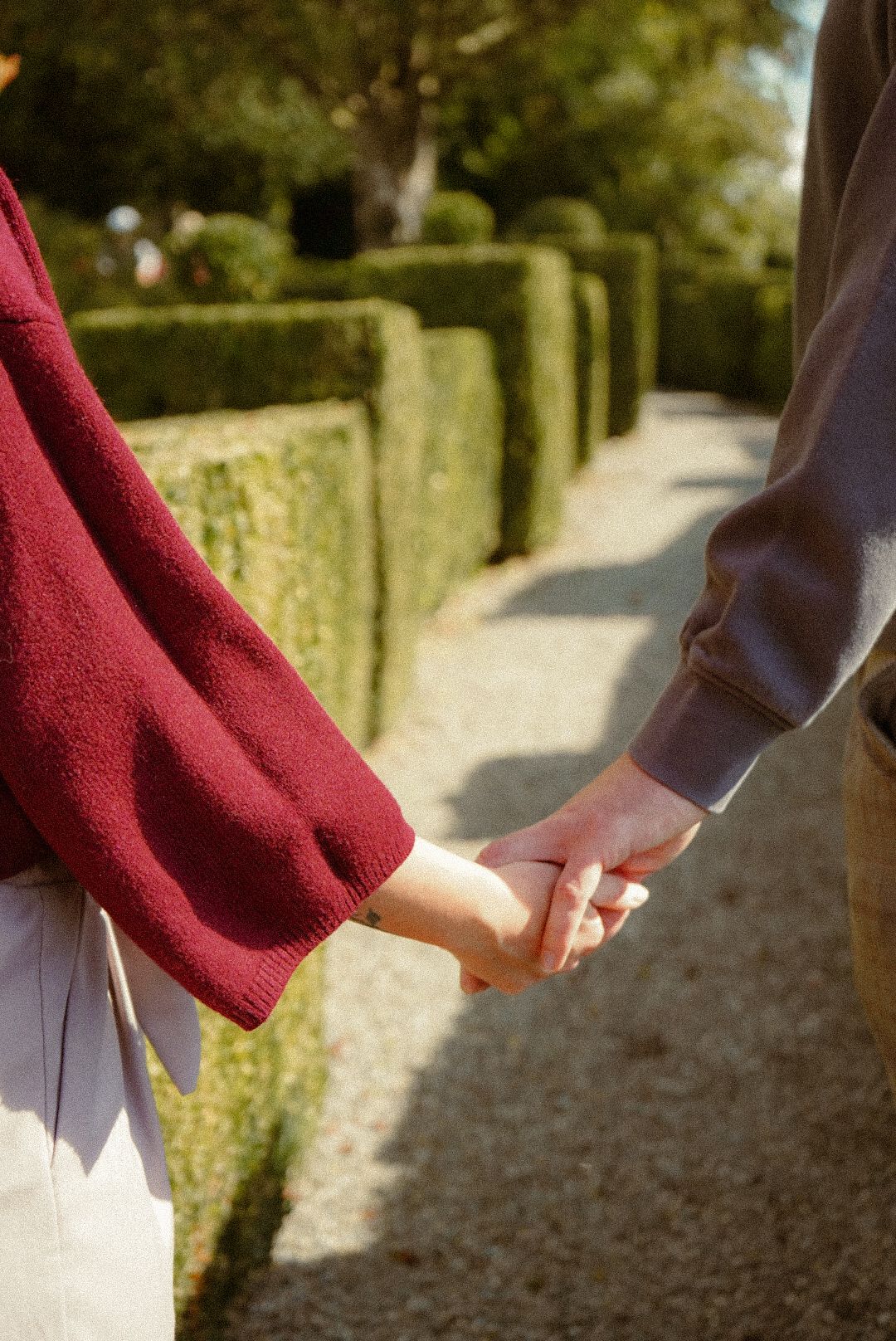 Two people holding hands while walking along a garden path with manicured hedges.